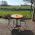 Compacte horecatafel voor buiten met warm houten tafelblad, twee groene rasterstoelen en een natuurlijke bamboeschutting op de achtergrond.