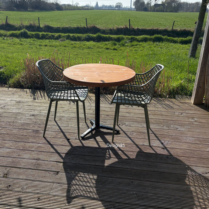 Kleine ronde terrastafel met zwart metalen poot en houten blad, geplaatst tussen twee groene kunststof stoelen in een zonnige tuin.
