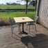 Industriële bartafel met houten blad op een terras in de tuin, omringd door planten en moderne stoelen.