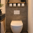 Industriële WC rolhouder Stijlvol van zwart staal met houten plank en vier toiletrollen, aan de muur gemonteerd boven een modern wit toilet in een sfeervolle toiletruimte.